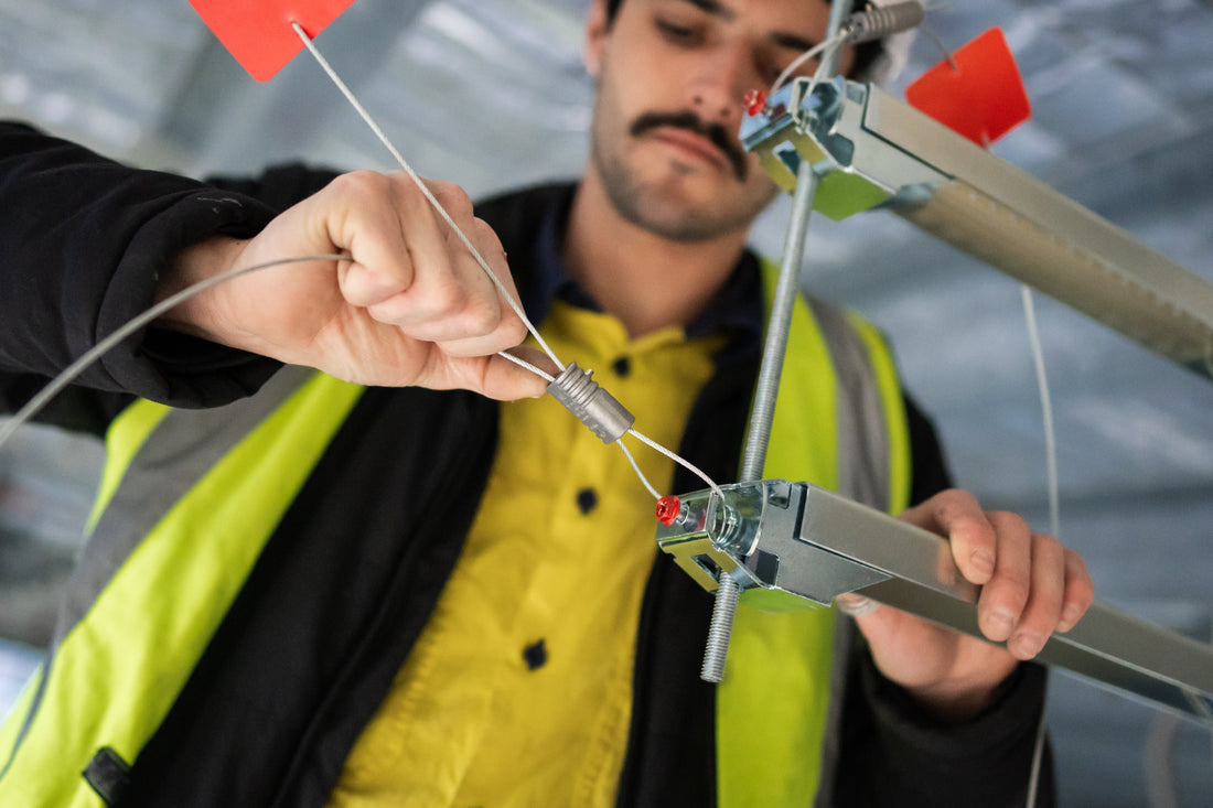 A worker wearing a yellow safety vest and black jacket installs a cable using a trapeze support adaptor in a construction setting.
