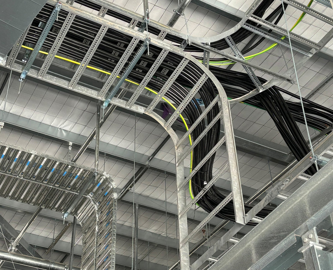 A close-up view of overhead cable trays with multiple bends, supporting electrical cables in a large industrial building.